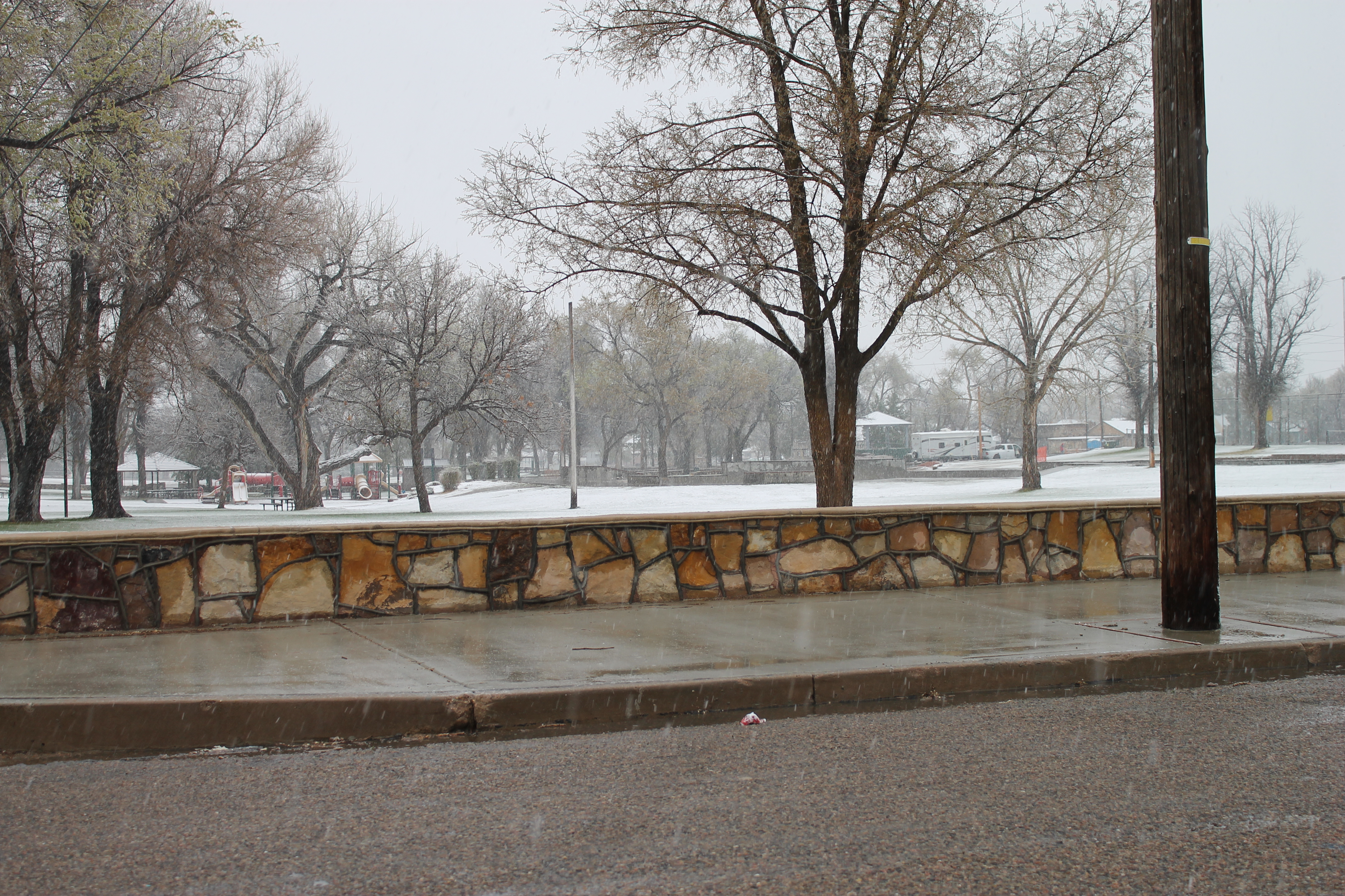 A photo of La Junta City Park Wall, a stone wall a couple of feet high set back from the road. Snow is on the ground.